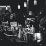 Black and white photo capturing the lively atmosphere in a bar with people socializing and a busy bartender serving drinks.