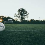 Home 7 Soccer ball on field at sunrise, serene landscape with goalpost in background.