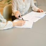 Home 7 woman signing on white printer paper beside woman about to touch the documents